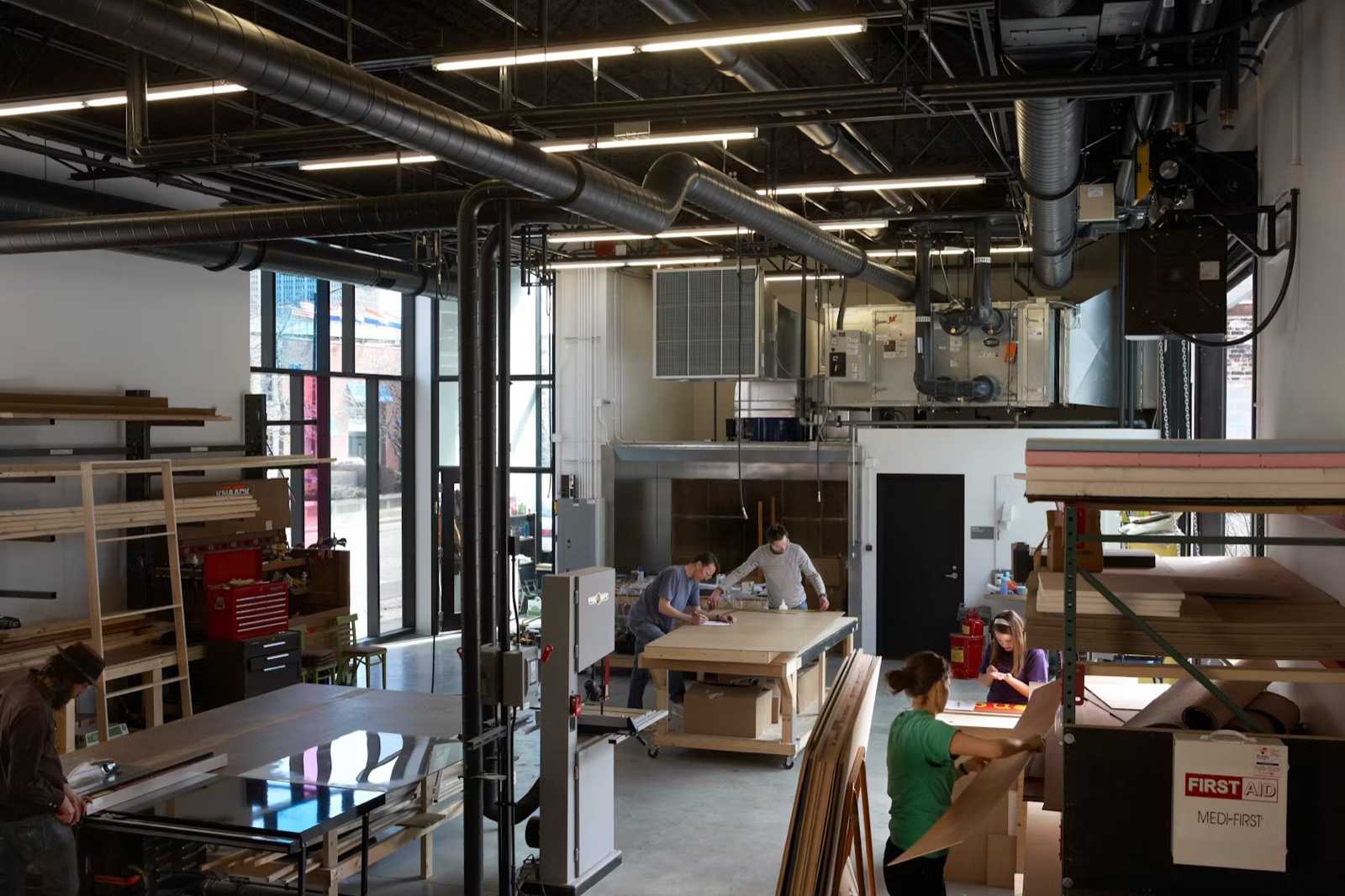 Wide view of a working fabrication lab — makers at work tables under industrial ductwork