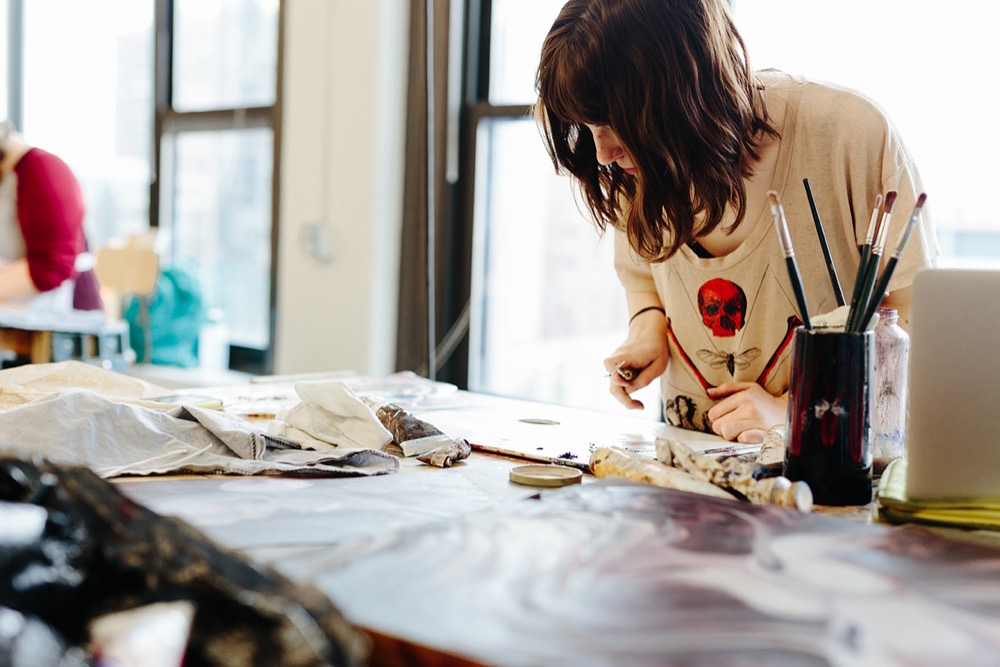 Artist working at a cluttered studio table in warm window light
